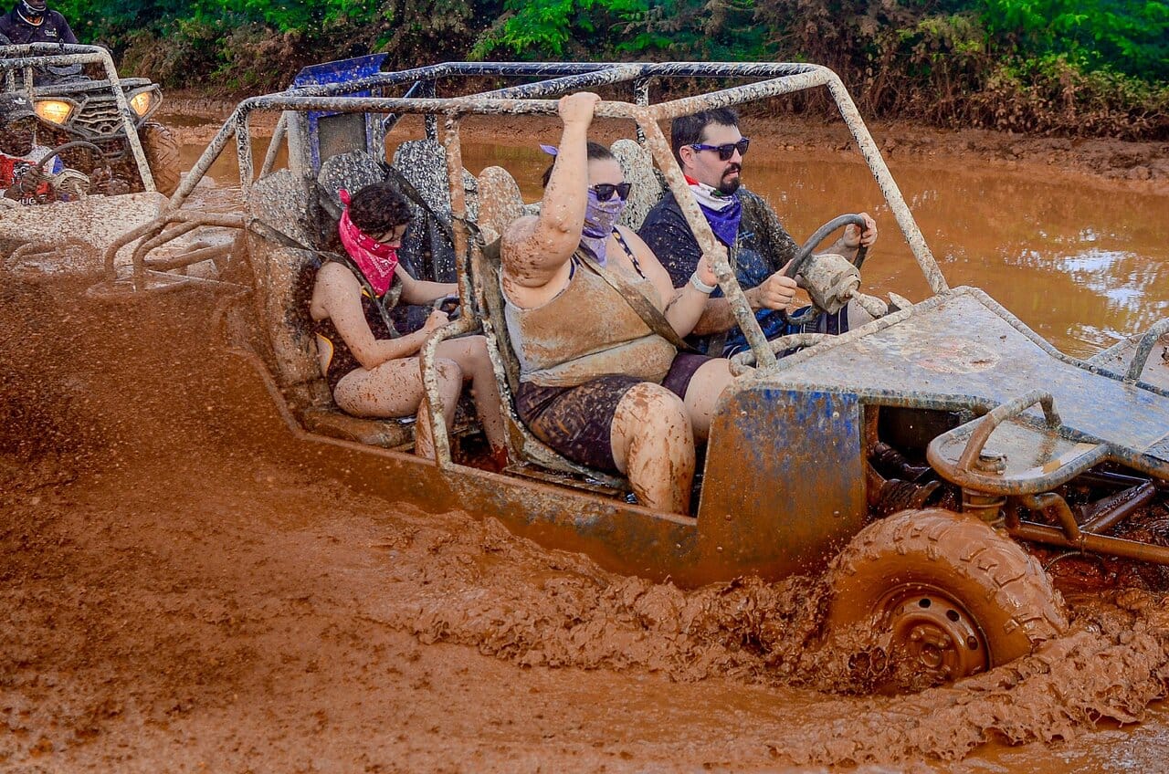 Three mud-splattered tourists in a buggy crossing a brown puddle, with another buggy behind them