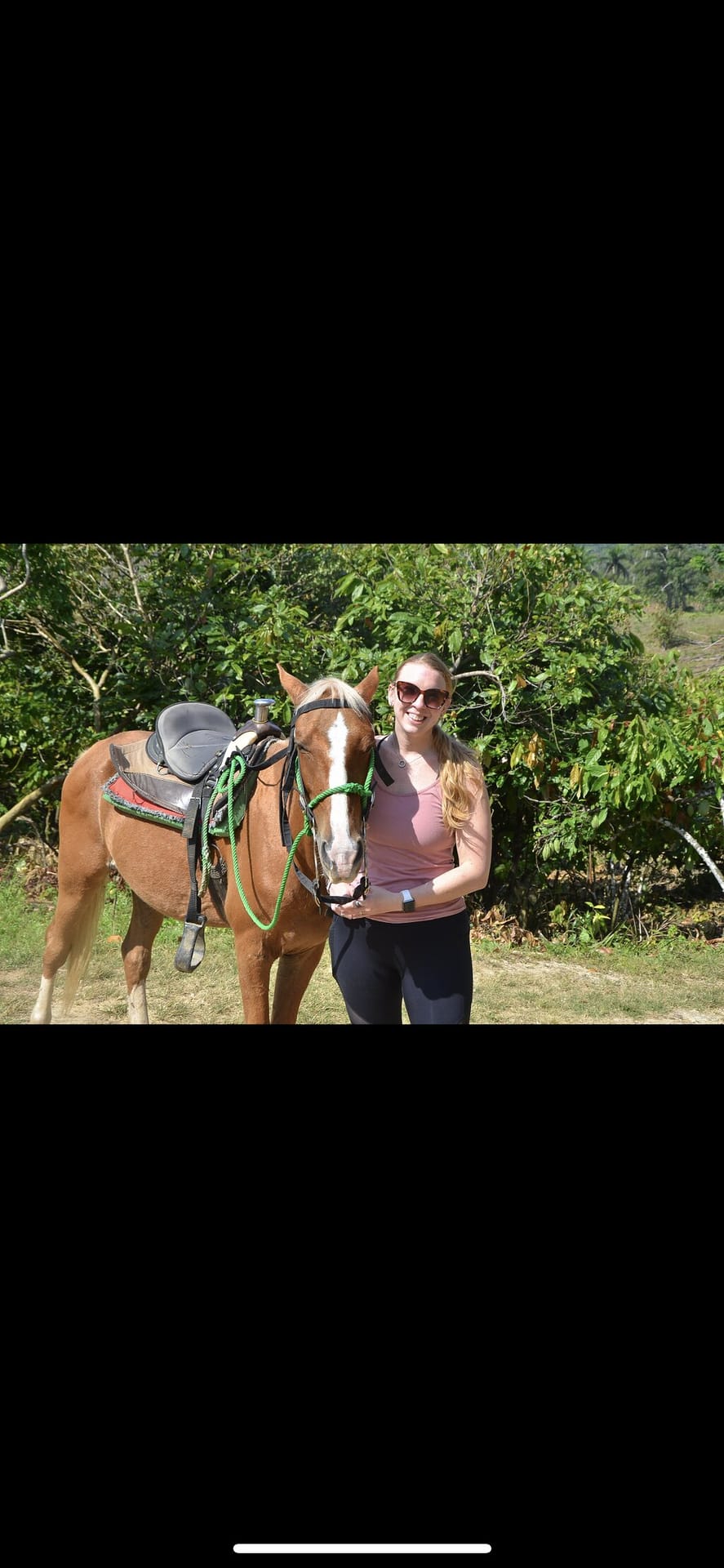 Tourist posing next to a saddled chestnut horse in a countryside setting with leafy trees