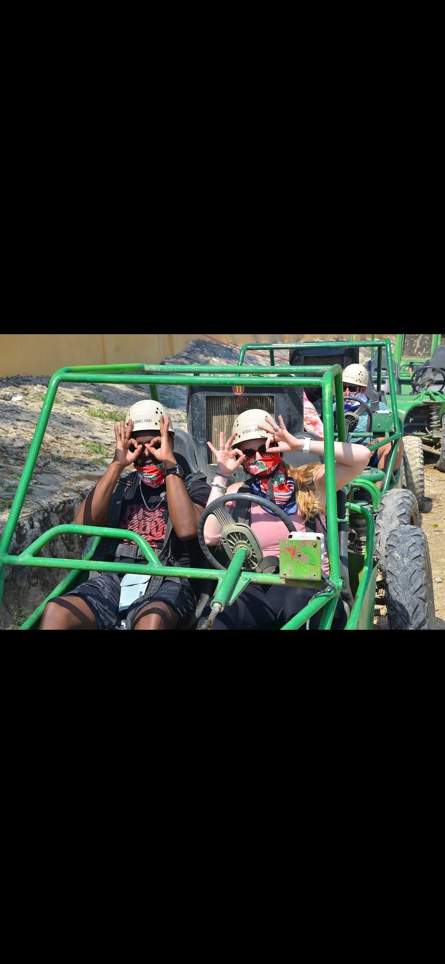 Two tourists in white helmets and bandanas making goggles with their hands inside a green buggy
