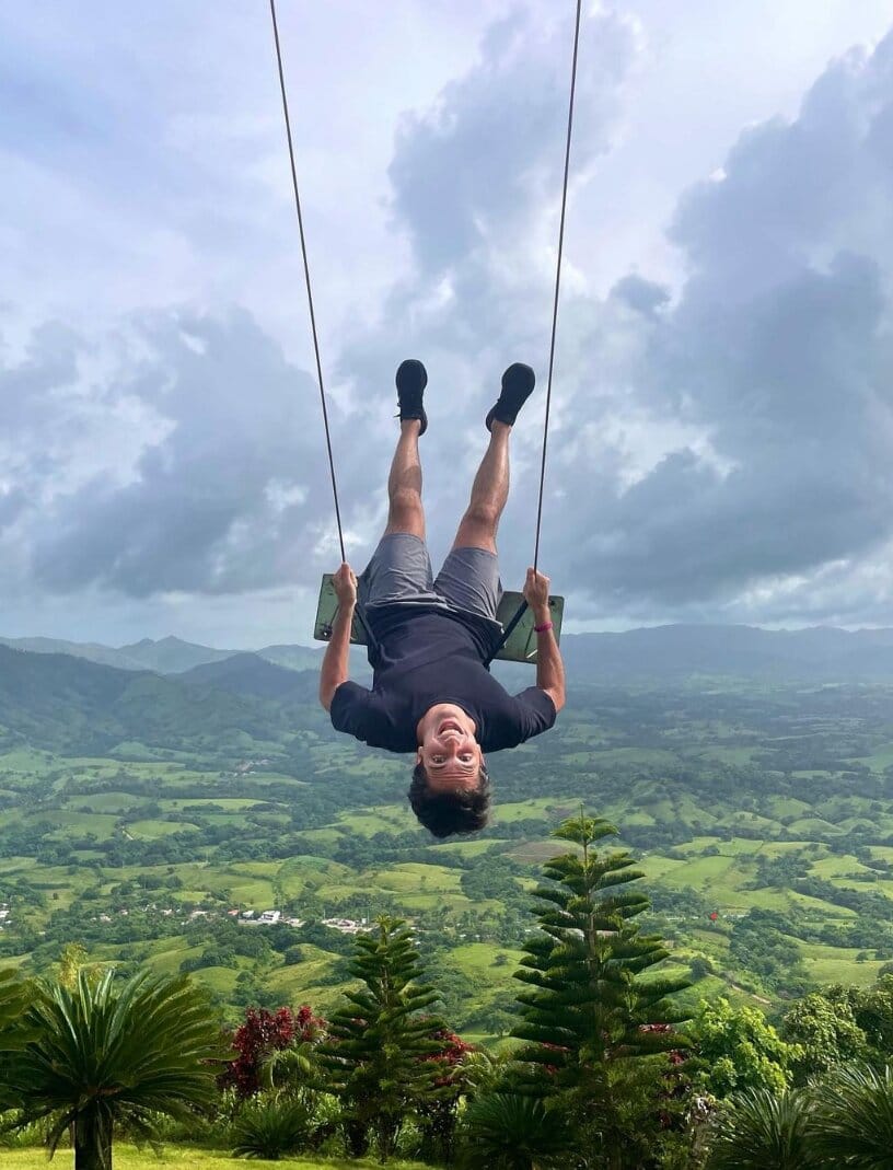 Tourist hanging upside down on a giant swing, with a green valley and mountains under a cloudy sky