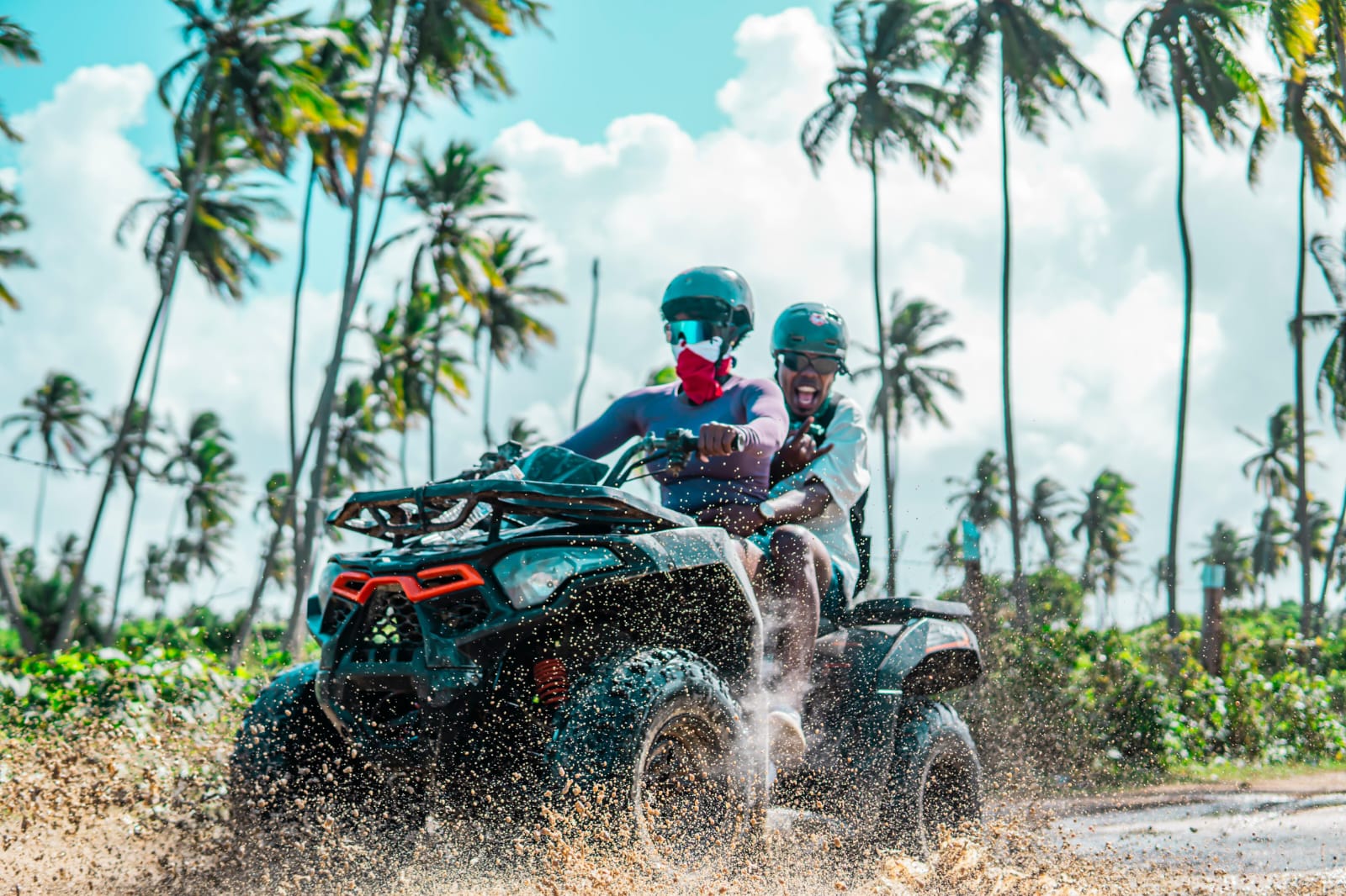 Two tourists in helmets on an ATV kicking up sand and mud on a trail surrounded by palm trees