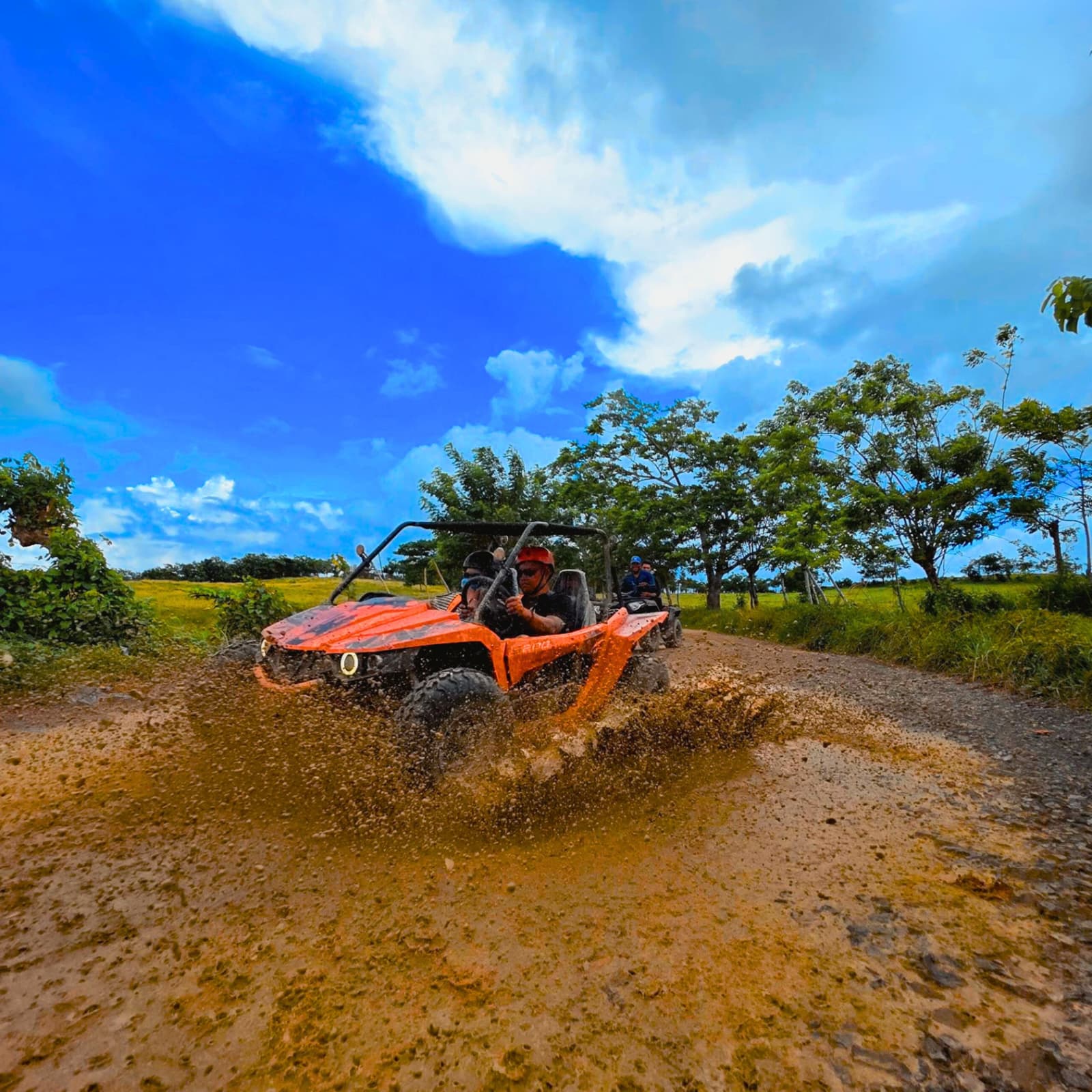 Orange buggy splashing mud as it crosses a puddle on a dirt road between green fields